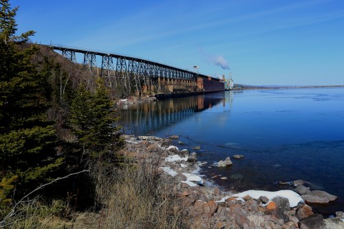 A tranquil river scene with a bridge, industrial buildings, and trees along the shore under a clear blue sky.