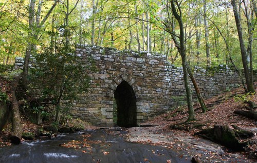 A stone arch bridge surrounded by trees and foliage, with a stream flowing in the foreground.