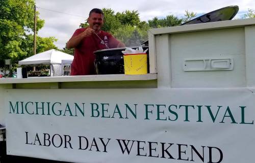 A man stirs a large pot at the Michigan Bean Festival, with a sign promoting Labor Day weekend in the background.