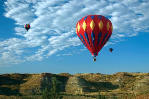 Colorful hot air balloons float over rolling hills under a blue sky with fluffy clouds.