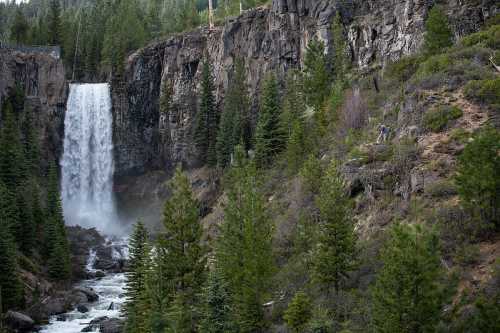 A waterfall cascades down rocky cliffs, surrounded by lush green trees and a flowing river below.