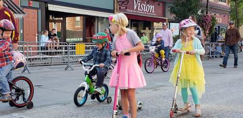 Children riding scooters and bikes in a festive street scene, with spectators watching from the sidelines.