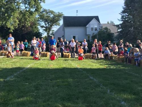 A crowd watches children participating in a fun outdoor event, sitting on hay bales in a grassy field.