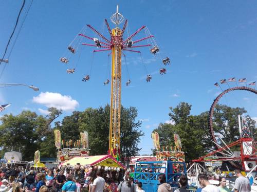 A vibrant carnival scene with a tall swing ride, colorful booths, and a crowd enjoying the festivities under a blue sky.