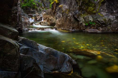 A serene river flows through rocky terrain, surrounded by lush greenery and smooth stones beneath the water's surface.