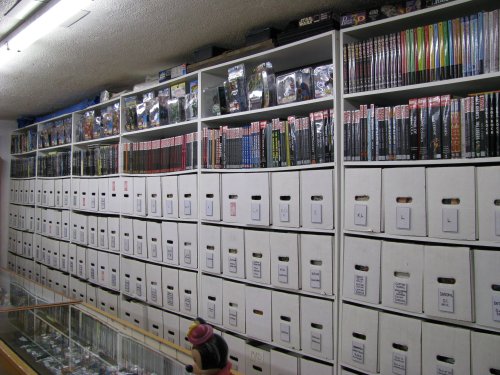 A wall of shelves filled with comic books and neatly organized boxes, with a display case in the foreground.