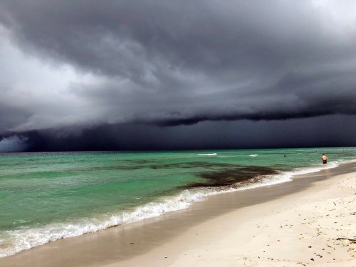 A beach scene with dark storm clouds looming over turquoise waters and a person walking along the shore.