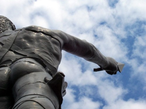 A close-up of a statue's back, with an outstretched arm pointing towards a cloudy sky.
