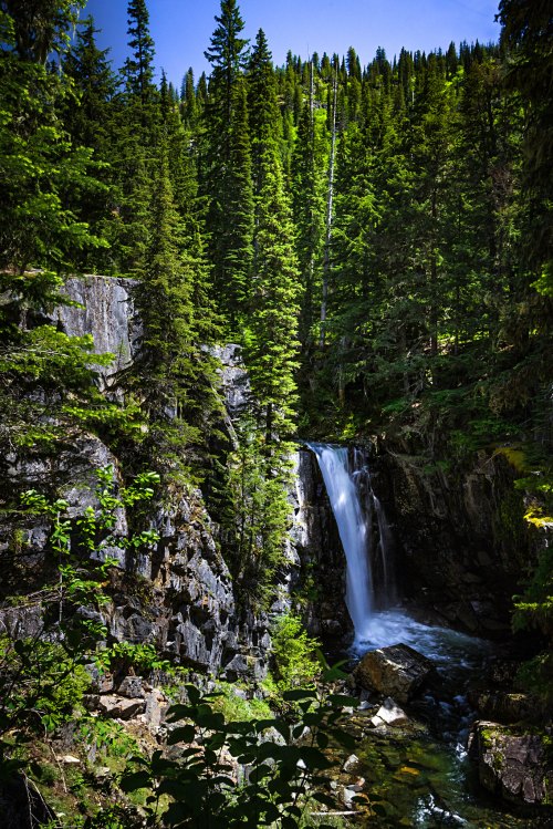 A serene waterfall cascades through lush green trees and rocky terrain under a clear blue sky.