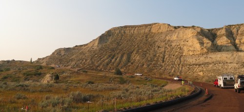 A winding road leads towards a large, rocky hill with a clear sky above and vehicles parked nearby.