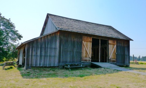 A rustic wooden barn with open doors, set in a grassy area under a clear blue sky.