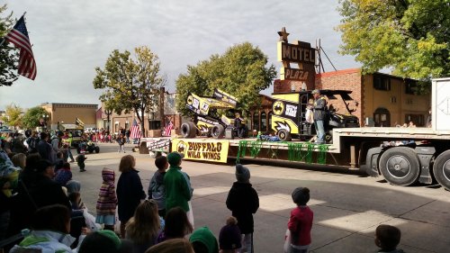 A parade float featuring a race car and Buffalo Wild Wings banner, with spectators watching on a sunny day.