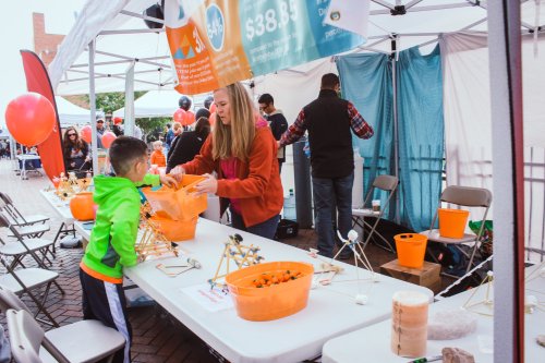 A woman assists a child at a science activity booth with colorful materials and structures on a table.