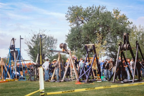 A crowd gathers around large wooden catapults at an outdoor event, with trees and blue sky in the background.