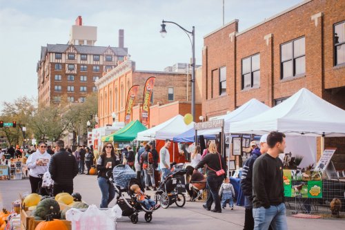 A bustling street market with vendors, tents, and people shopping, surrounded by autumn decorations and buildings.