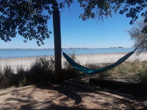A tranquil beach scene featuring a hammock tied between trees, with calm water and distant islands in the background.