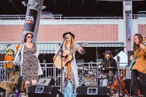 A band performs on stage, featuring a singer with an acoustic guitar and three musicians playing various instruments.