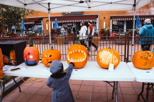 A child reaches for a carved pumpkin at a festive outdoor market with people and colorful decorations in the background.