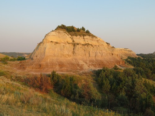 A large, layered rock formation rises above a grassy landscape under a clear sky during sunset.