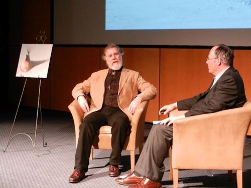 Two men sit in armchairs on stage, discussing a presentation with a photo displayed on an easel beside them.