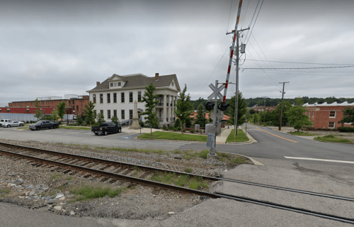 A view of a street intersection with a railroad crossing, featuring a large building and parked cars nearby.