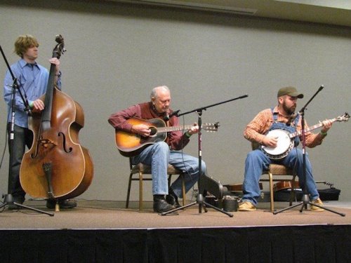 Three musicians perform on stage: one with a double bass, one with an acoustic guitar, and one with a banjo.
