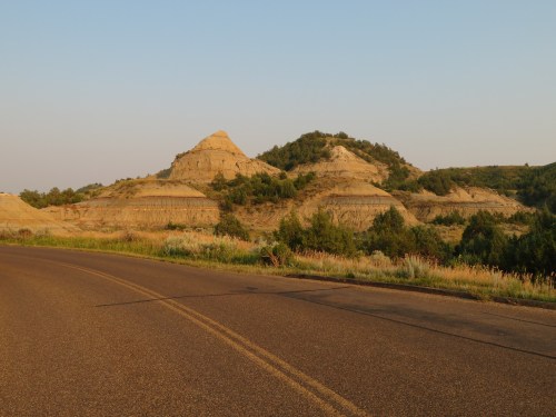 A scenic view of rolling hills and layered rock formations beside a paved road under a clear sky.
