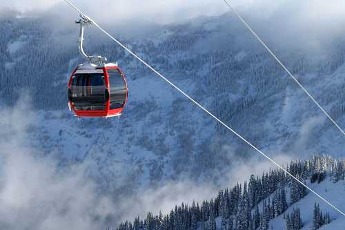 A red gondola glides above snowy mountains, surrounded by clouds and evergreen trees.