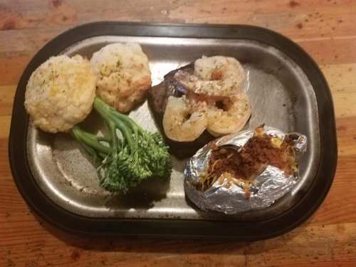 A metal tray with two baked shrimp, broccoli, two breaded muffins, and a baked potato wrapped in foil.