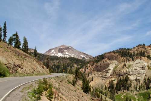 A winding road leads through a mountainous landscape with snow-capped peaks and lush greenery under a blue sky.