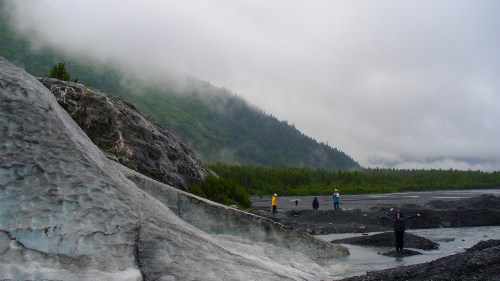 People exploring a glacier landscape with misty mountains and a forest in the background.
