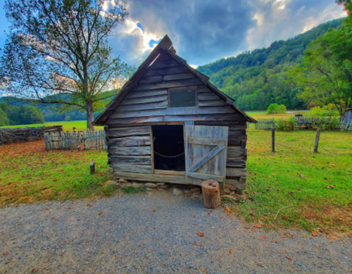 A rustic wooden cabin with a partially open door, surrounded by greenery and hills under a cloudy sky.