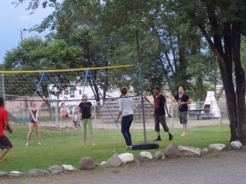 A group of children and adults play in a park with swings and a tire swing under trees.