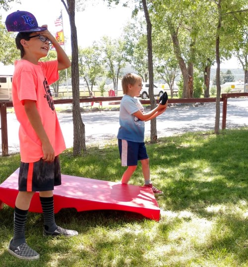 Two children play outside on a sunny day, one wearing a cap and the other holding a disc, near a red cornhole board.