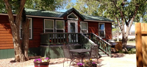 A cozy wooden cabin with a green roof, surrounded by trees, featuring a front porch and outdoor seating area.