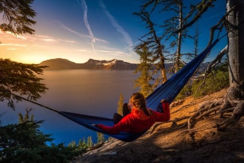 A person relaxes in a hammock by a serene lake at sunset, surrounded by trees and mountains.