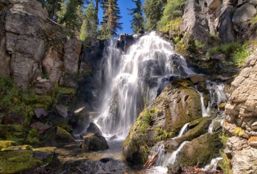 A serene waterfall cascading over rocky terrain, surrounded by lush greenery and tall trees under a clear blue sky.