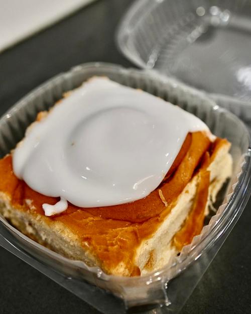 A close-up of a cinnamon roll topped with white icing in a clear plastic container.