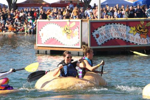 Two women paddle a giant pumpkin in a festive water event, surrounded by a cheering crowd and colorful banners.