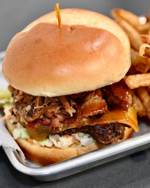 A close-up of a pulled pork burger with cheese, pickles, and coleslaw, served with a side of crispy fries.