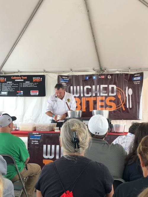 A chef demonstrates cooking techniques at a culinary tent during the Wicked Bites event, with an audience watching.