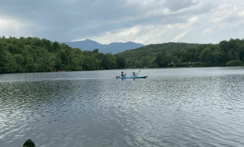 Two people kayak on a calm lake surrounded by green trees and mountains under a cloudy sky.