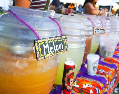 Colorful beverage dispensers labeled with flavors like "Melon" and "Pepino" on a festive table at a market.