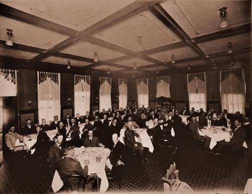 A large group of people dining in a vintage restaurant, seated at tables with white tablecloths and elegant decor.
