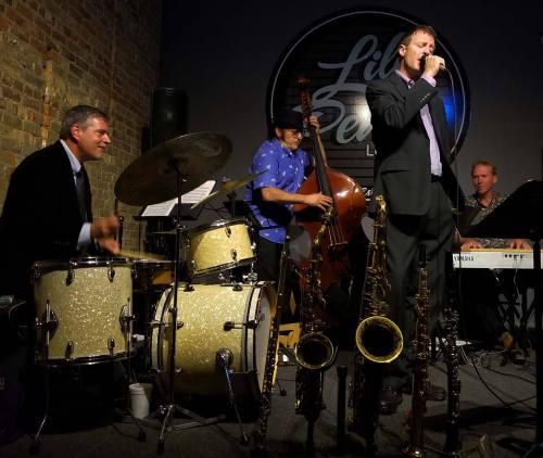 A jazz band performs on stage, featuring a vocalist, drummer, bassist, and keyboardist with saxophones in the foreground.