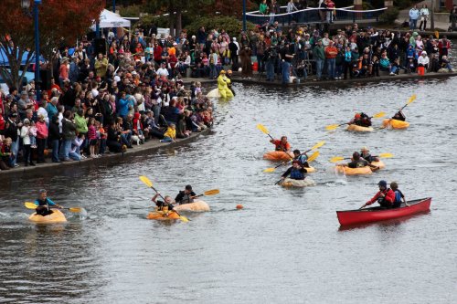 A crowd watches as participants paddle in kayaks and canoes on a river during a lively outdoor event.