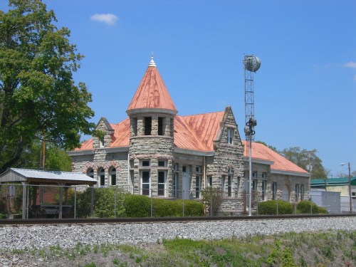 Historic stone building with a copper roof, surrounded by greenery and near a railway track under a clear blue sky.