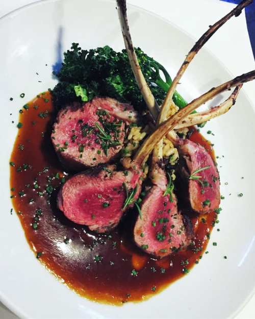A plate of lamb chops with a rich sauce, garnished with herbs and served with greens and a side of grains.