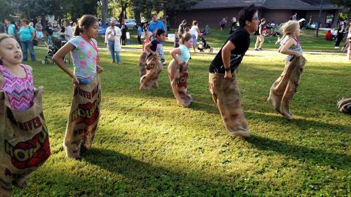 Children participate in a sack race on a grassy field during a community event, smiling and jumping in burlap sacks.