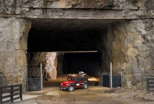 A red truck parked at the entrance of a large stone cave, with rocky walls and a dark interior visible.